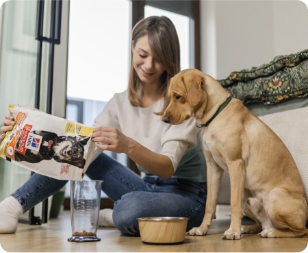 หญิงคนหนึ่งกำลังเทอาหารสัตว์เลี้ยงยี่ห้อ Hill's Pet Nutrition ลงในภาชนะตวง ในขณะที่สุนัขตัวหนึ่งมองดูอย่างตั้งใจ